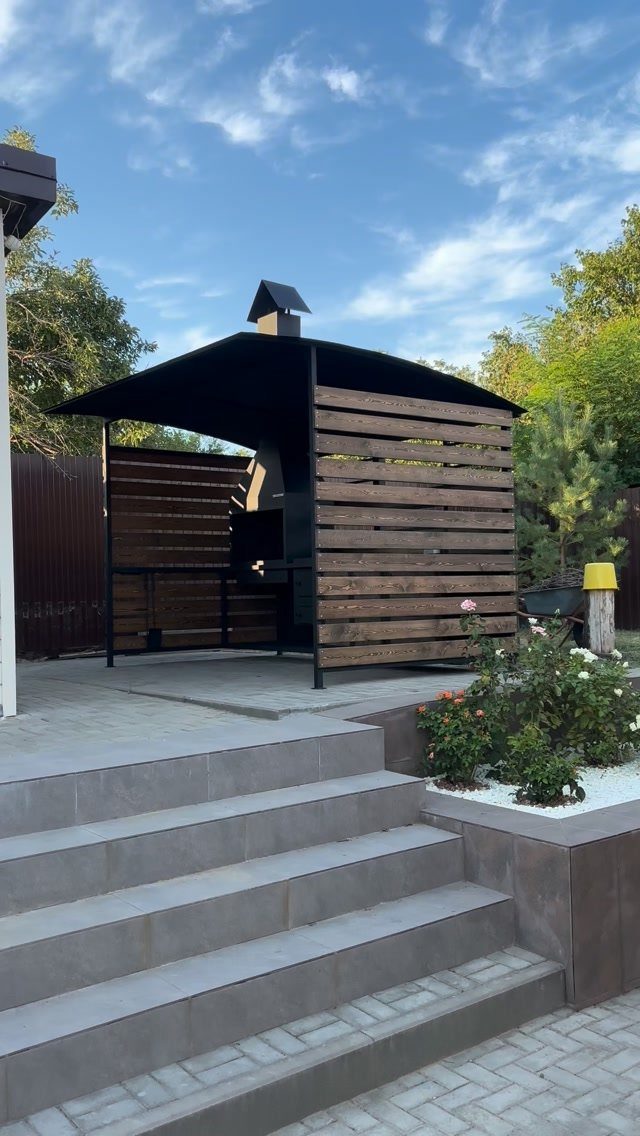 A modern black metal and wood slat outdoor BBQ shelter in a landscaped backyard.