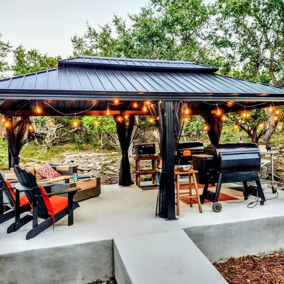 A black metal gazebo with string lights, a grill, and lounge seating on a concrete patio.