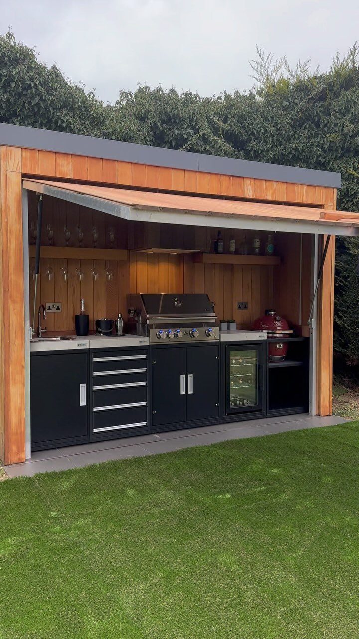 A wooden outdoor BBQ shelter featuring black cabinetry, a built-in grill, and a small refrigerator.