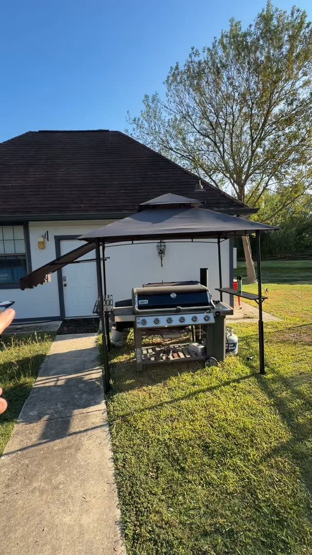 A black metal frame canopy shelter covering a stainless steel gas grill on a sunny lawn.