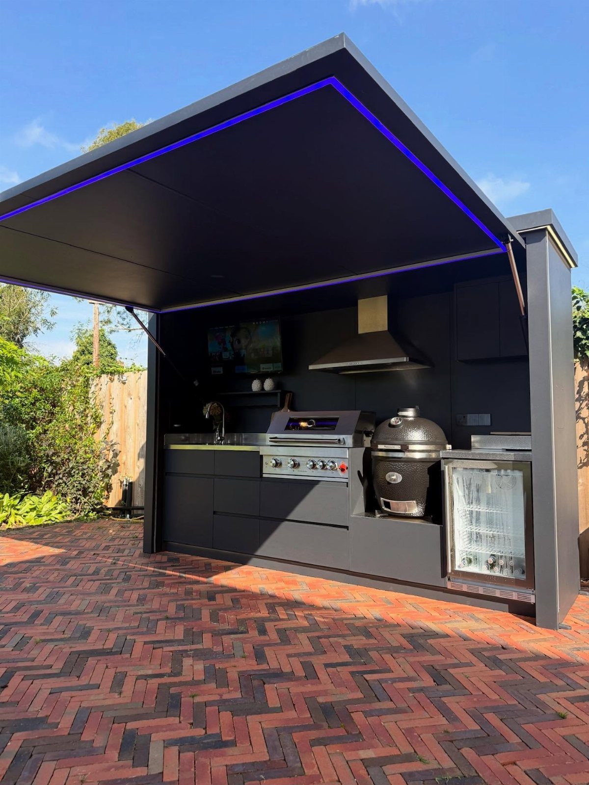 Modern dark grey outdoor kitchen shelter with blue LED lighting and built-in barbecue equipment.