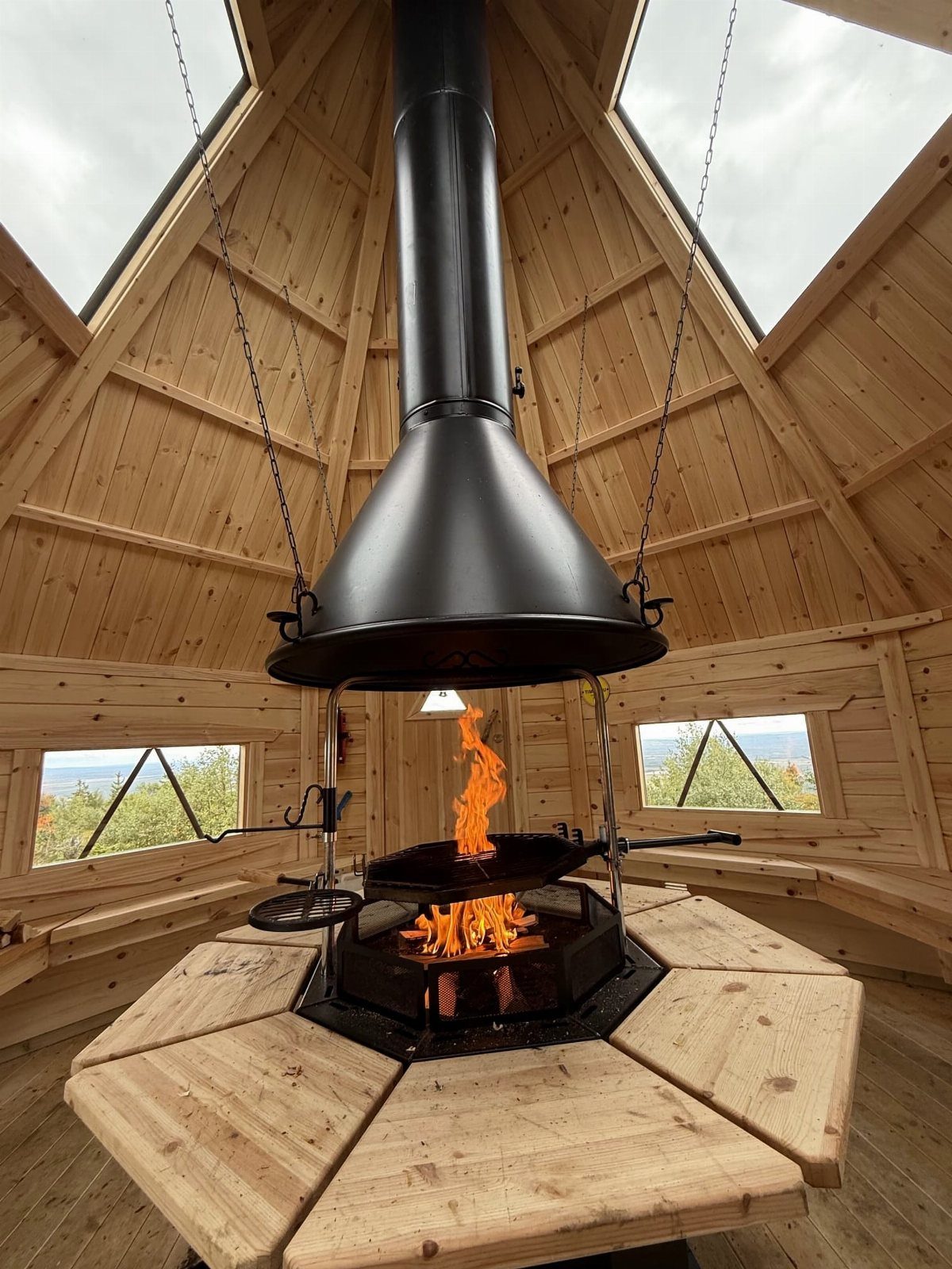 A black metal fireplace hood hangs over a burning fire pit inside a wooden hut.