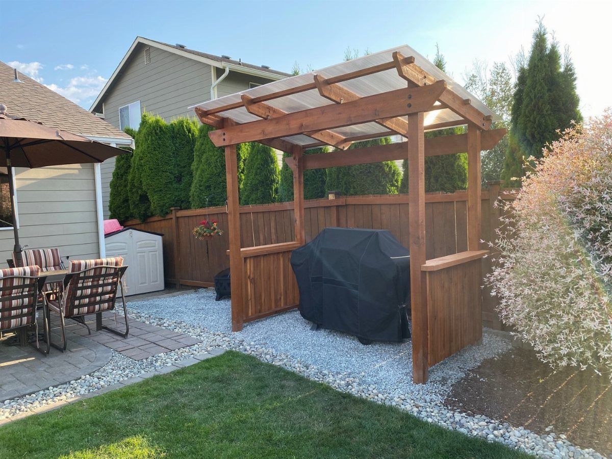 A wooden frame shelter with a translucent roof protecting a covered barbecue grill outdoors.