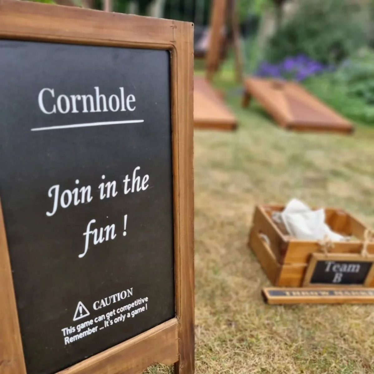 A wooden chalkboard sign promoting cornhole stands in front of cornhole boards on a lawn.