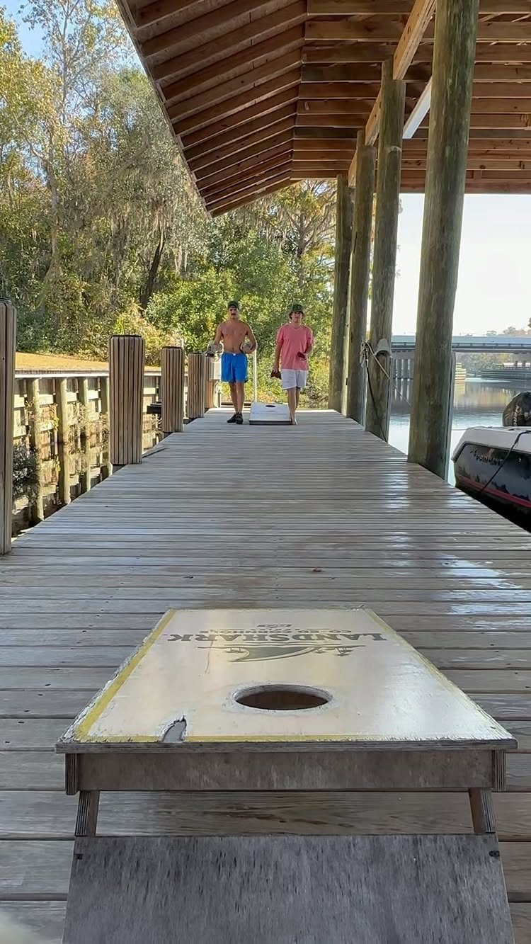 A cornhole board sits on a wooden dock with people walking in the background.