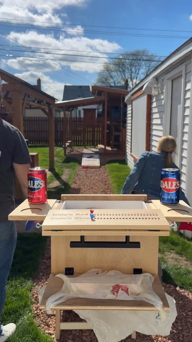 A wooden cornhole board station with soda cans on a gravel backyard path.