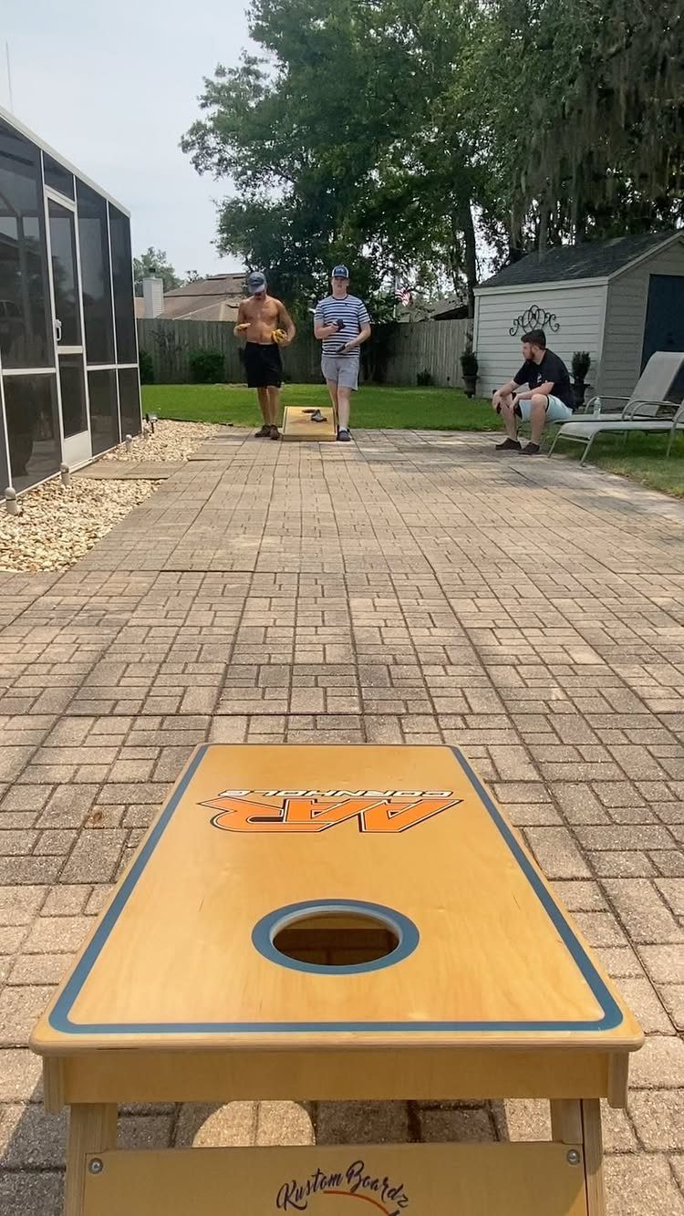 A wooden cornhole board sits on a stone patio with people playing in the background.