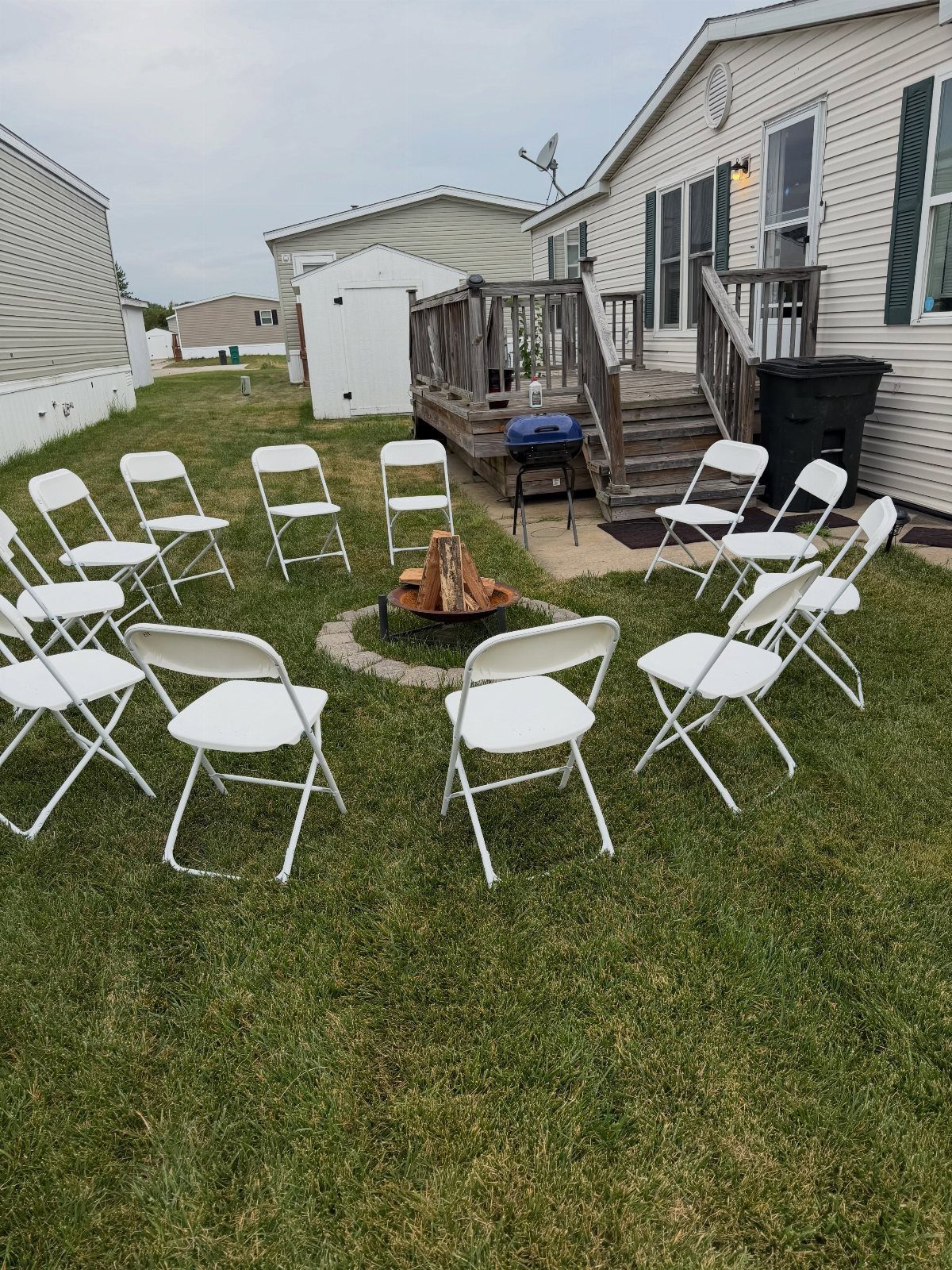 White folding chairs arranged in a circle on grass around a small burning fire pit.