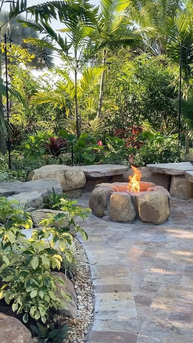 A stone fire pit with a bright flame on a paved patio surrounded by tropical greenery.