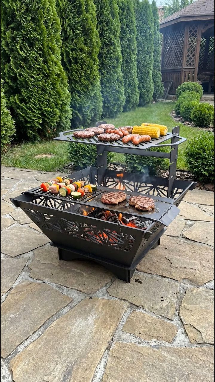 A two-tier metal fire pit grill cooking burgers, sausages, corn, and vegetable skewers outdoors.