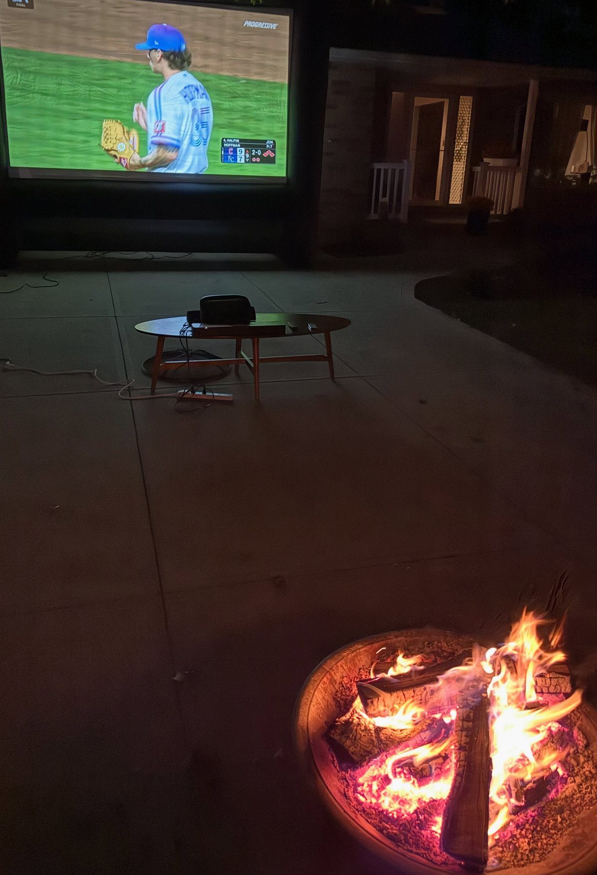 A glowing fire pit in the foreground with a large outdoor movie screen in the background.