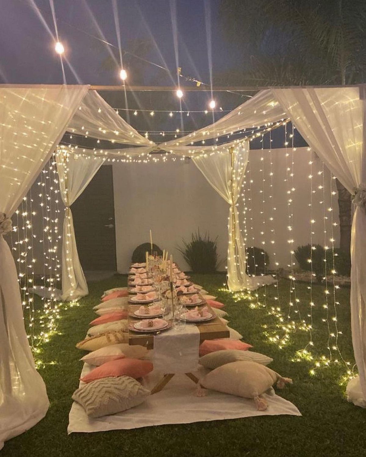 A low outdoor dining table set with cushions and fairy lights under white fabric draping.