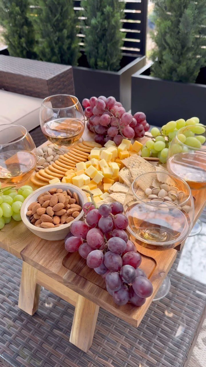 A wooden platter with grapes, cheese, crackers, almonds, and glasses of rose wine.