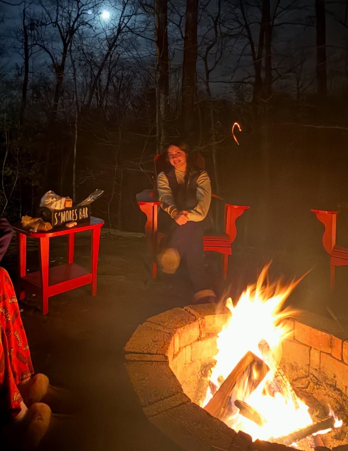 A roaring fire in a stone pit with a s'mores bar on a red table nearby.