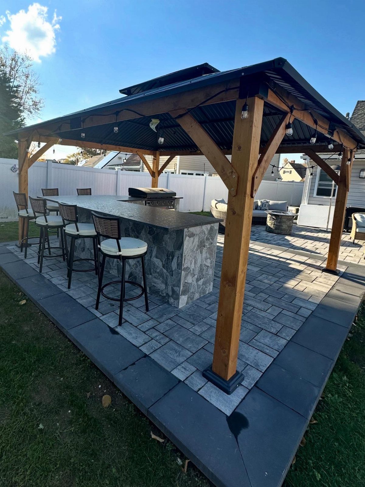 A wooden gazebo structure sheltering a stone outdoor bar with stools on a paved patio.