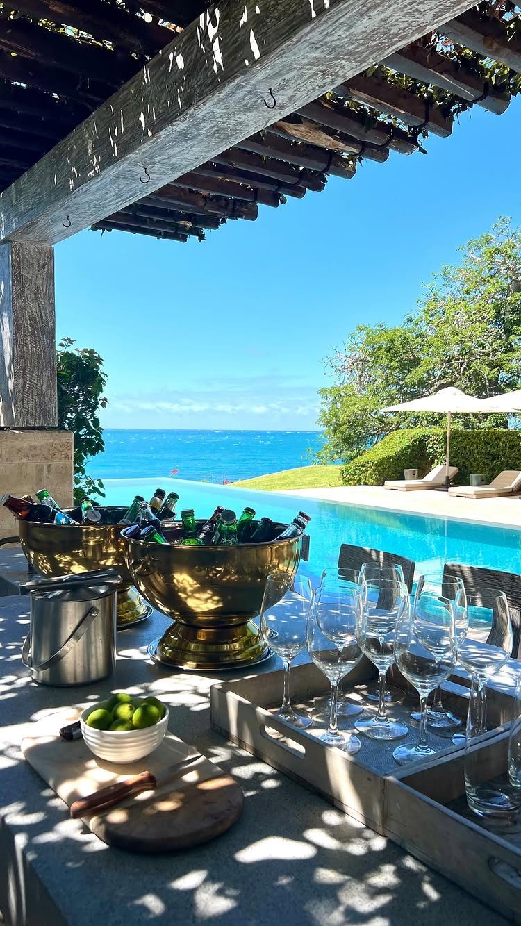 Gold ice buckets and glassware sit on a stone bar overlooking a pool and ocean.