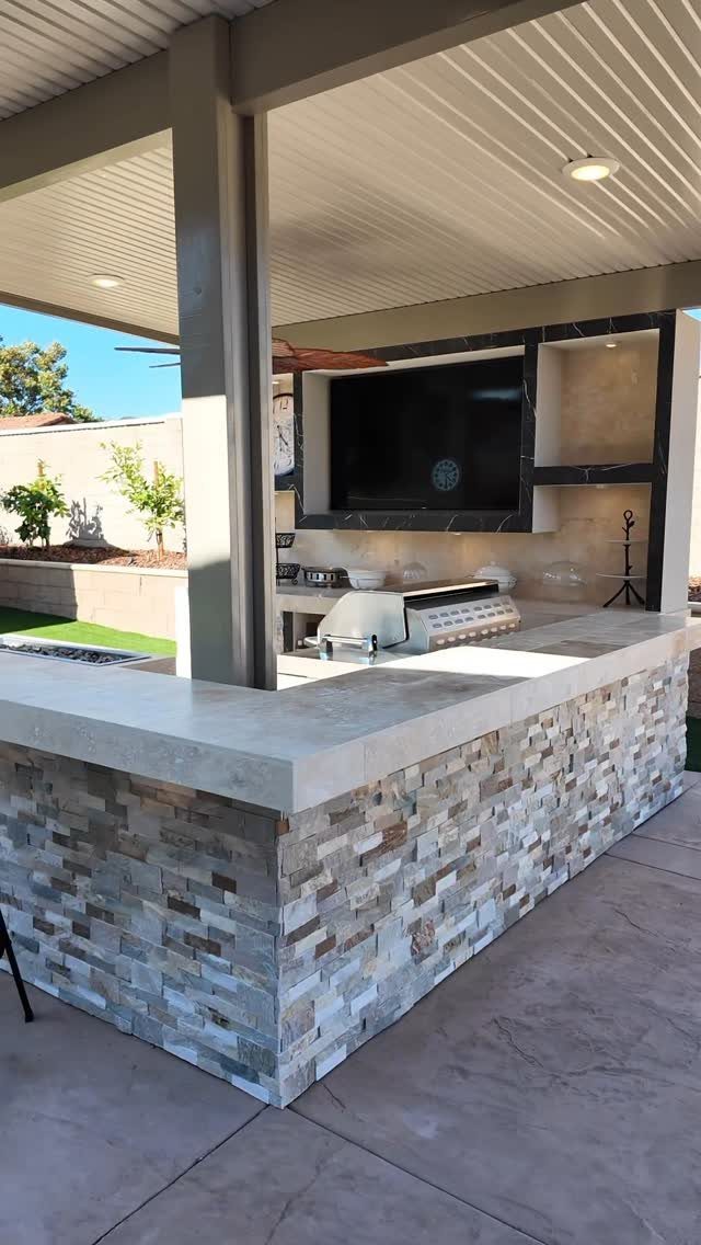 A stone-faced outdoor kitchen bar with a large countertop under a covered patio structure.