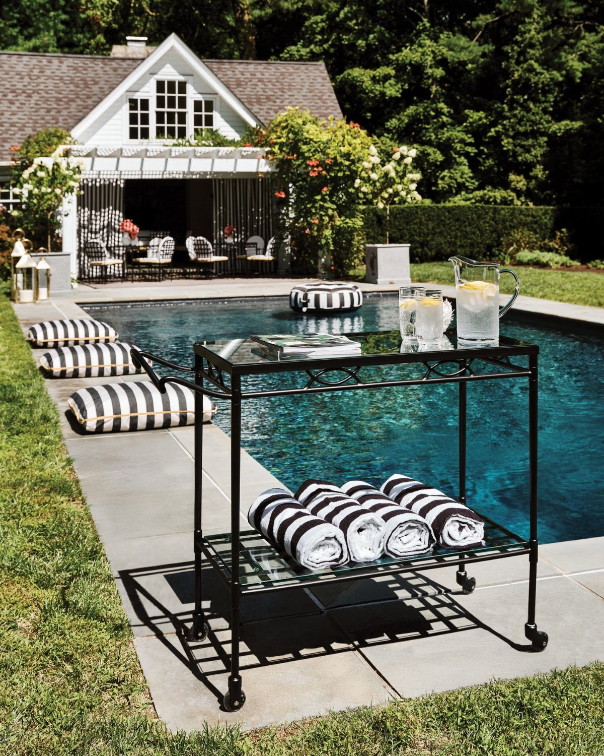 A black metal bar cart with lemonade and striped towels sits beside a backyard swimming pool.