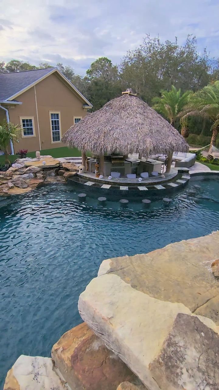 A thatched-roof tiki bar situated at the edge of a large swimming pool with tropical landscaping.
