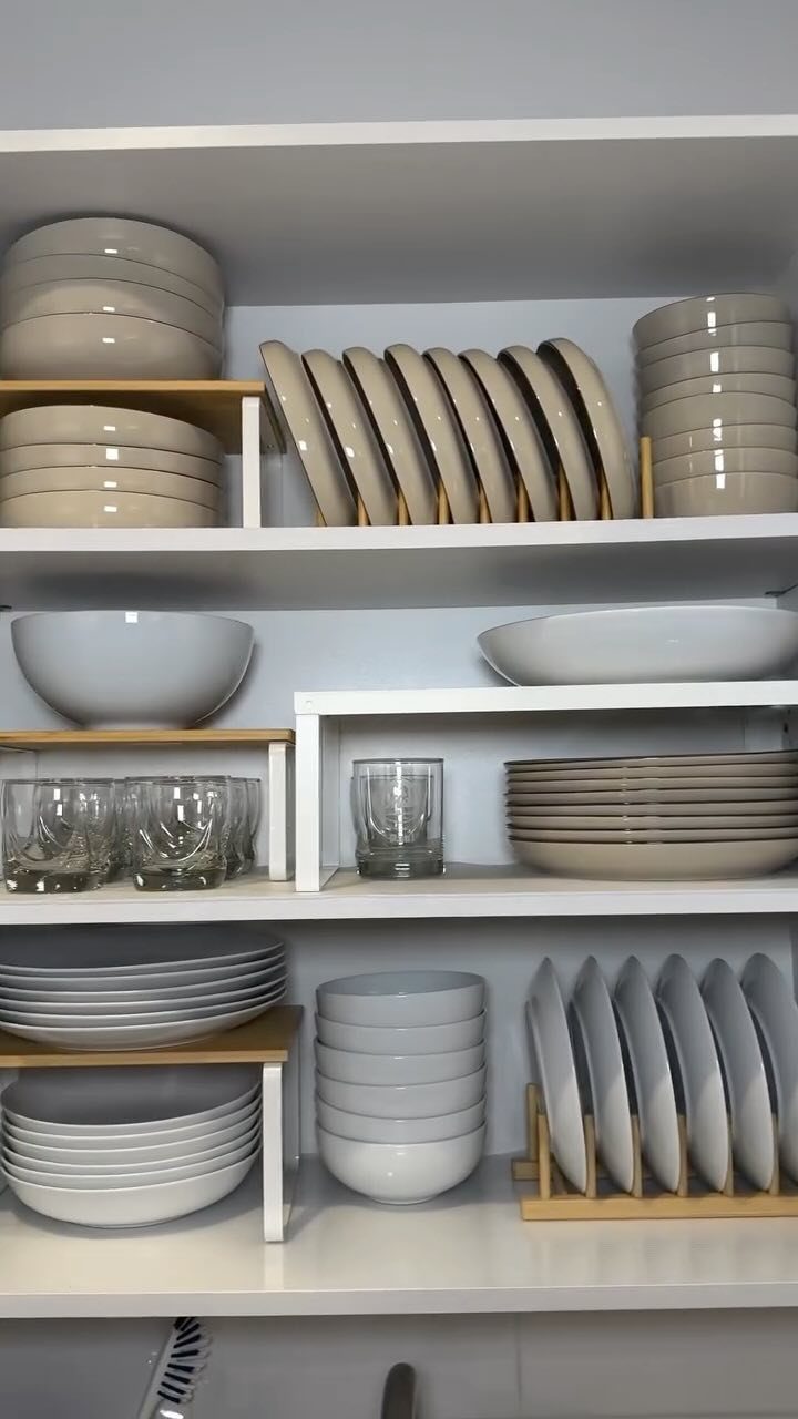 Stacks of white and beige plates and bowls organized on tiered shelving inside white cabinets.