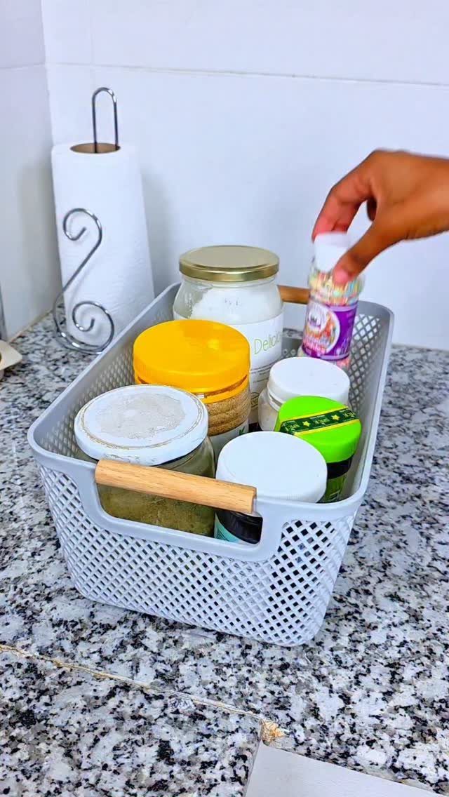 A hand reaches into a grey plastic basket filled with various spice jars on a granite countertop.