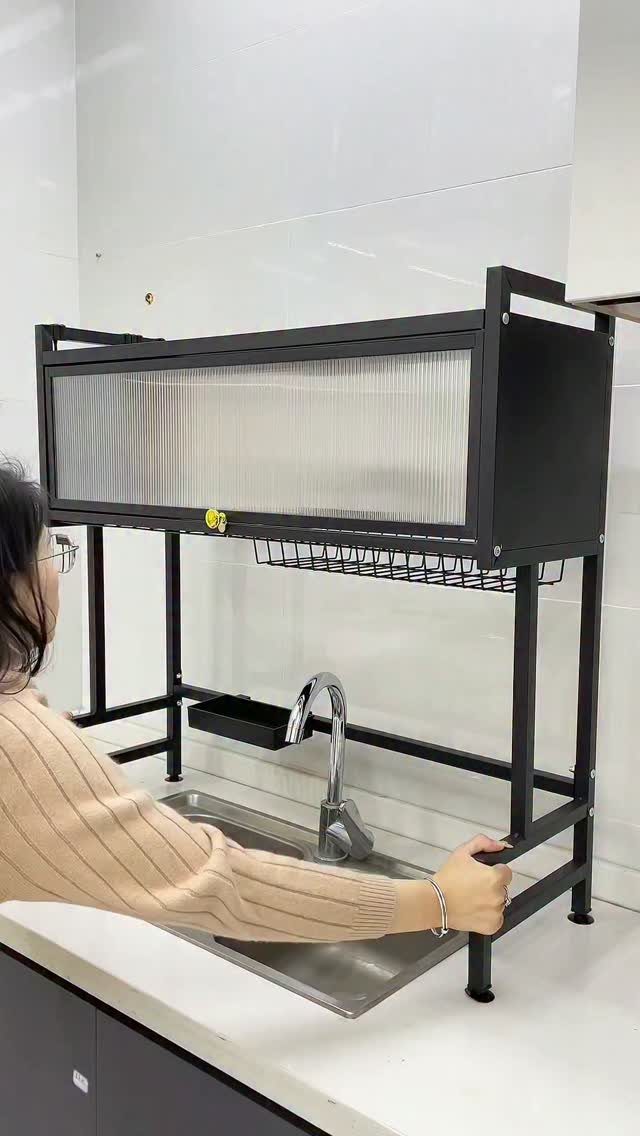 A person adjusts a black metal over-the-sink storage rack above a kitchen sink.