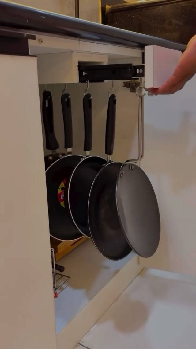 A hand pulls out a white cabinet drawer containing four hanging black frying pans.