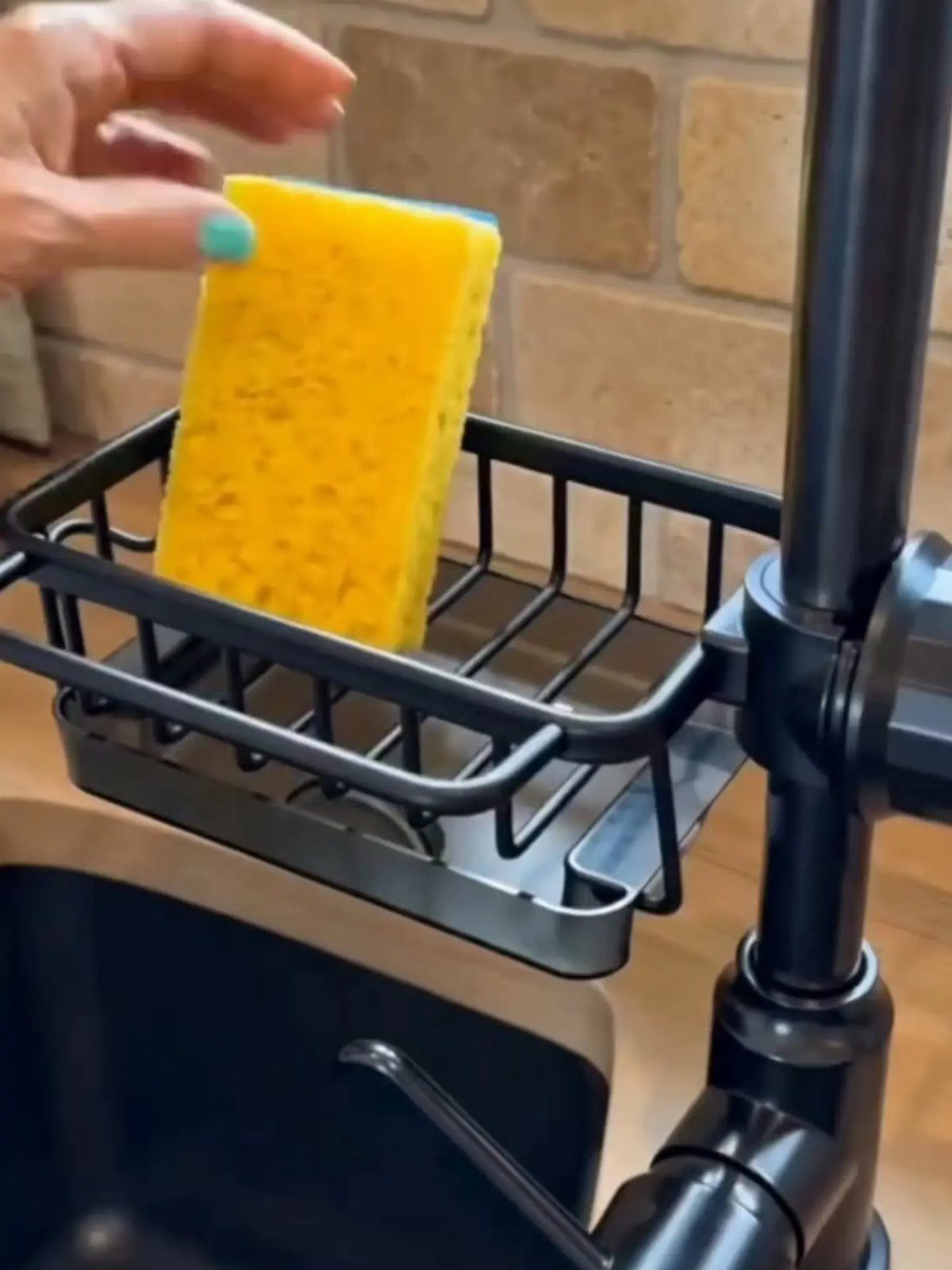 A hand placing a yellow sponge into a black wire rack organizer near a kitchen faucet.