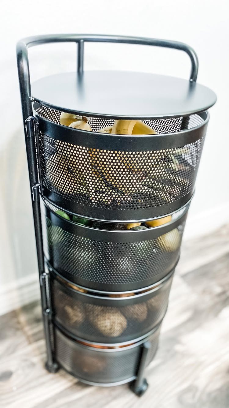 A black metal tiered basket stand filled with bananas and potatoes on a wooden floor.