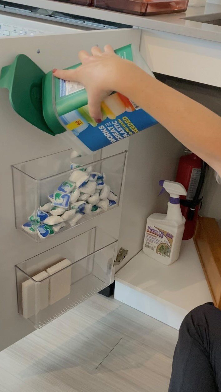 A hand reaches for plastic wrap stored in a clear organizer on a cabinet door.