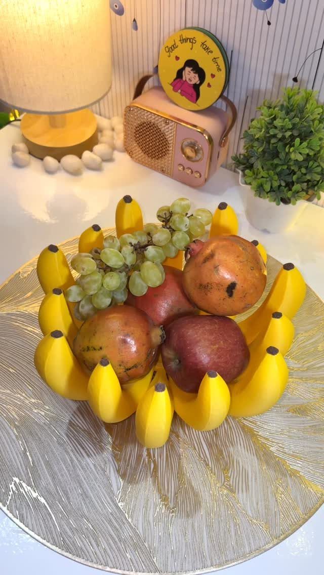 A gold banana-shaped bowl filled with grapes, pomegranates, and apples on a white surface.