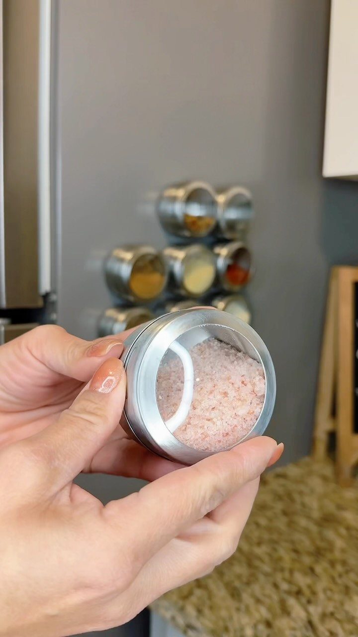 A person holding a small glass spice jar filled with pink salt in a kitchen.