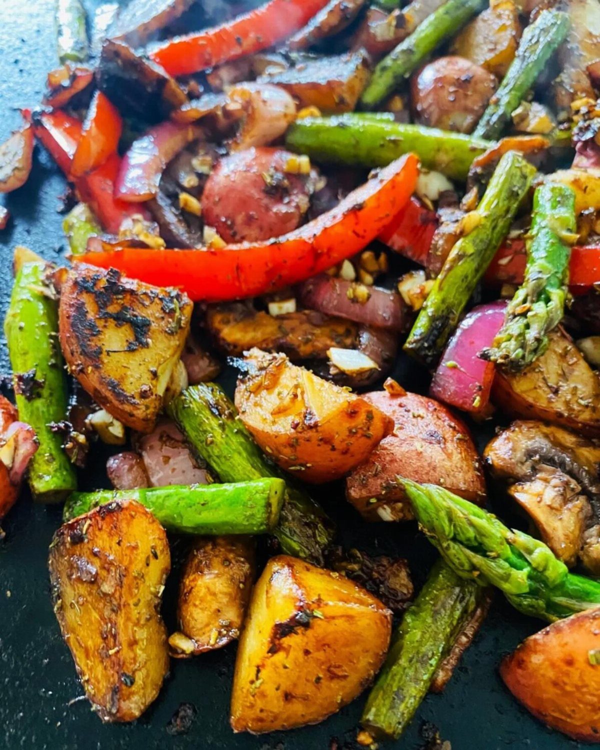Sautéed red peppers, asparagus, and seasoned potatoes cooking on a dark griddle surface.