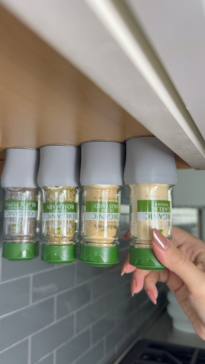Four spice jars hanging from the underside of a wooden kitchen cabinet above grey tiles.