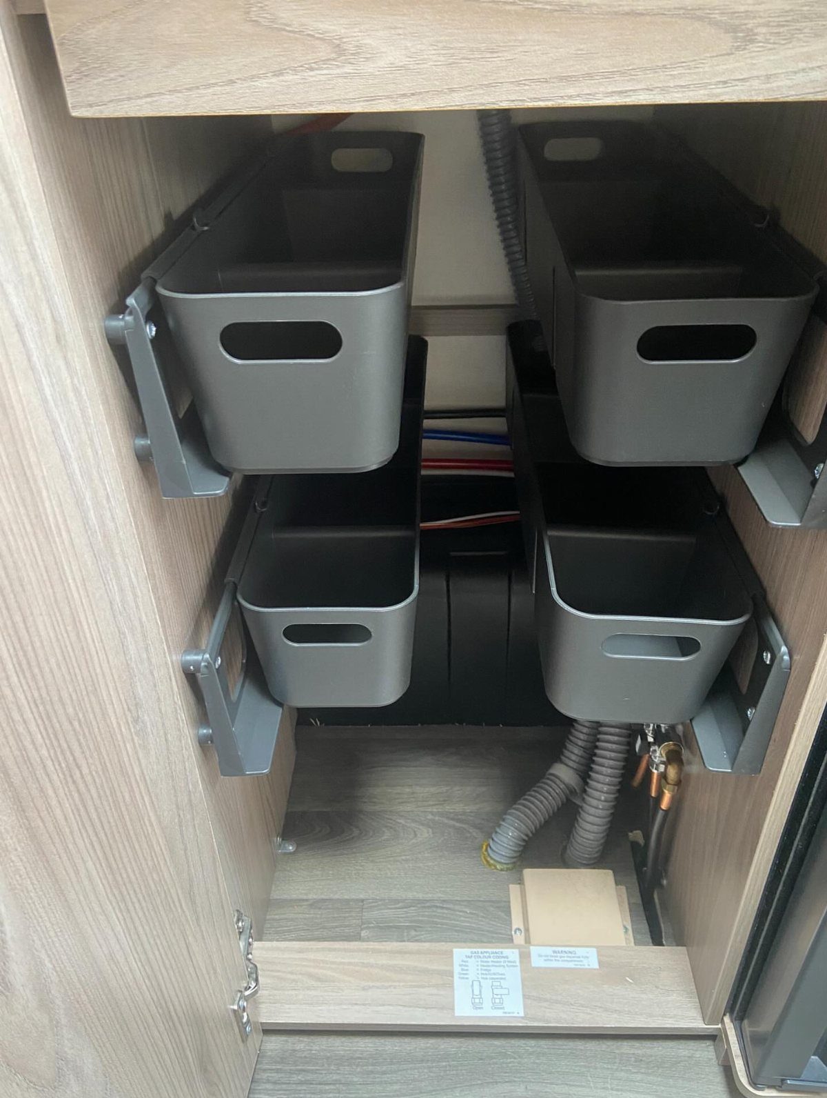 Four grey storage bins mounted on brackets inside a wooden camper cabinet compartment.