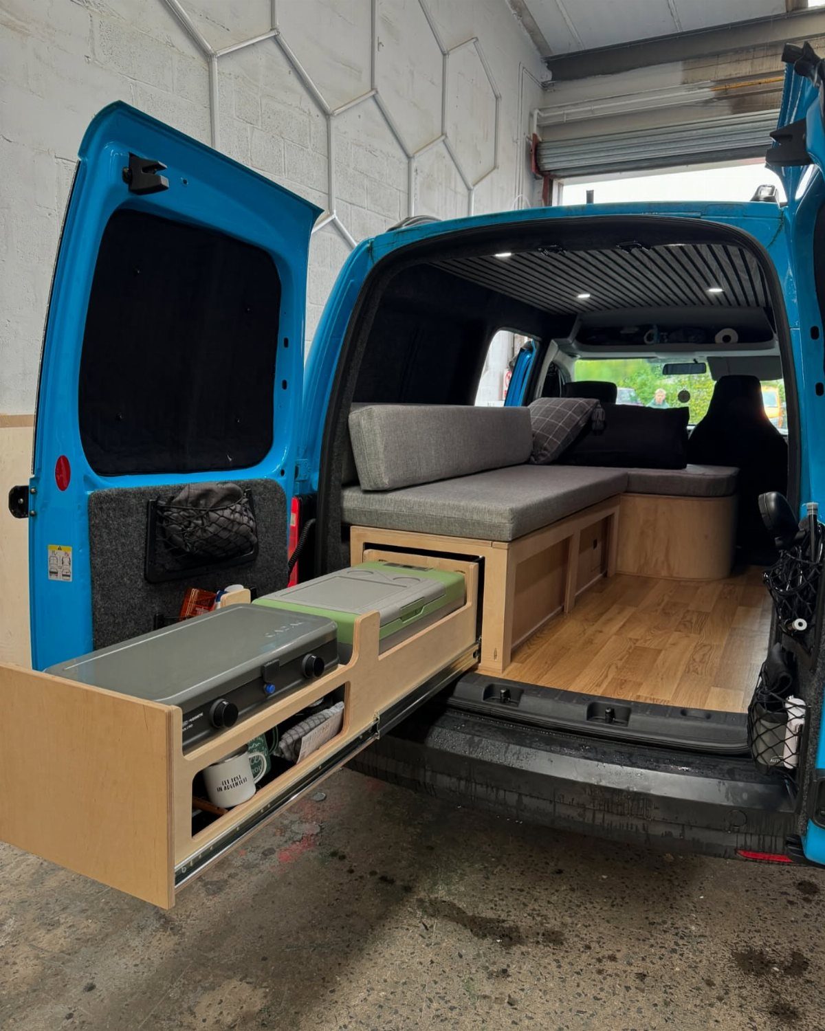 A wooden slide-out drawer containing a portable stove is extended from a blue camper van.