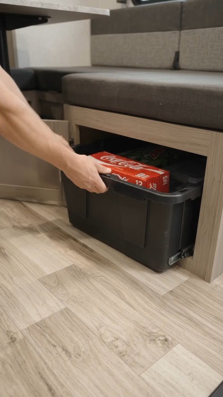 A hand pulling a black plastic storage bin from under a camper bench seat.