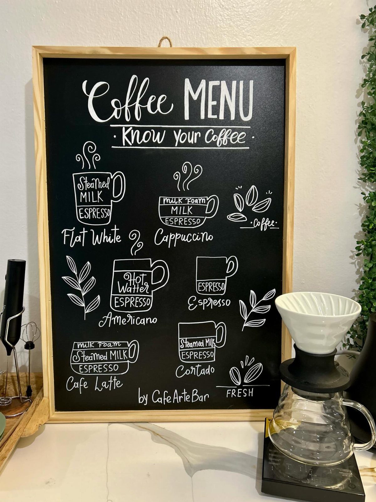 A chalkboard coffee menu with handwritten drink descriptions sitting on a white kitchen counter.