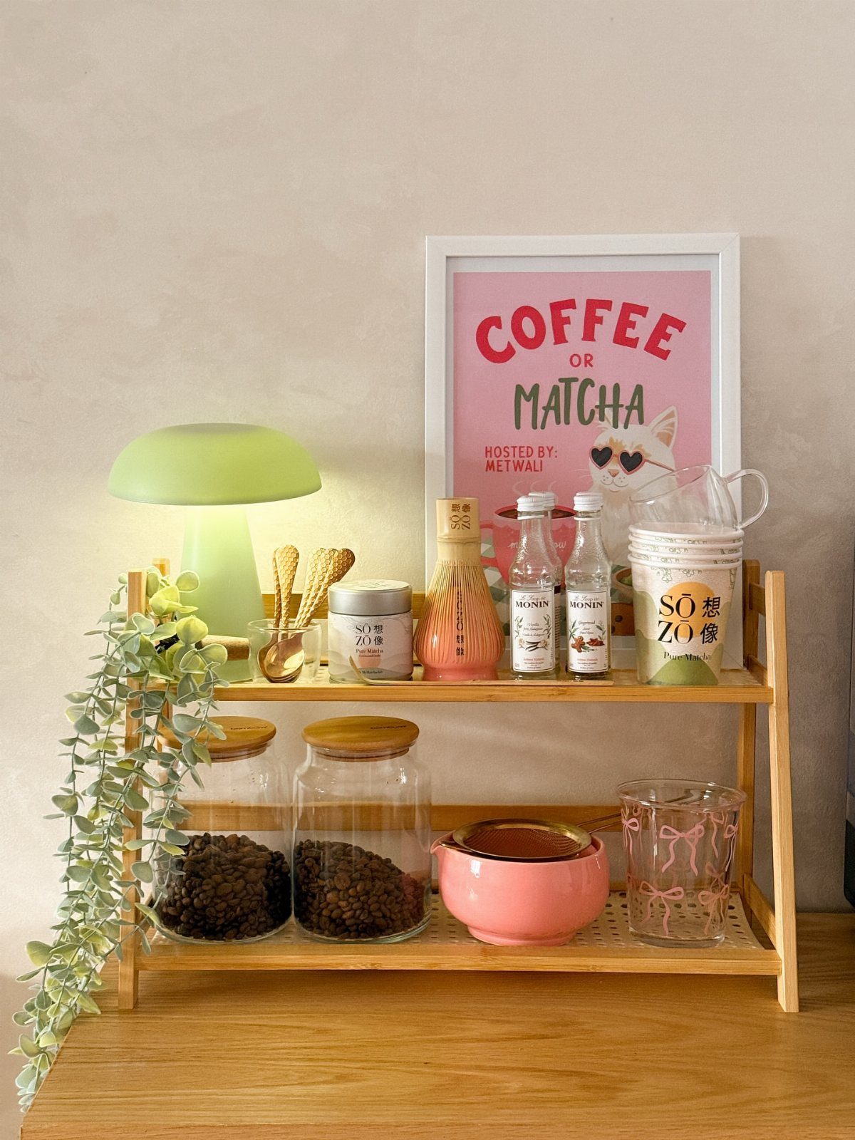 A wooden two-tier shelf holding coffee beans, bottles, and a green lamp with a trailing plant.