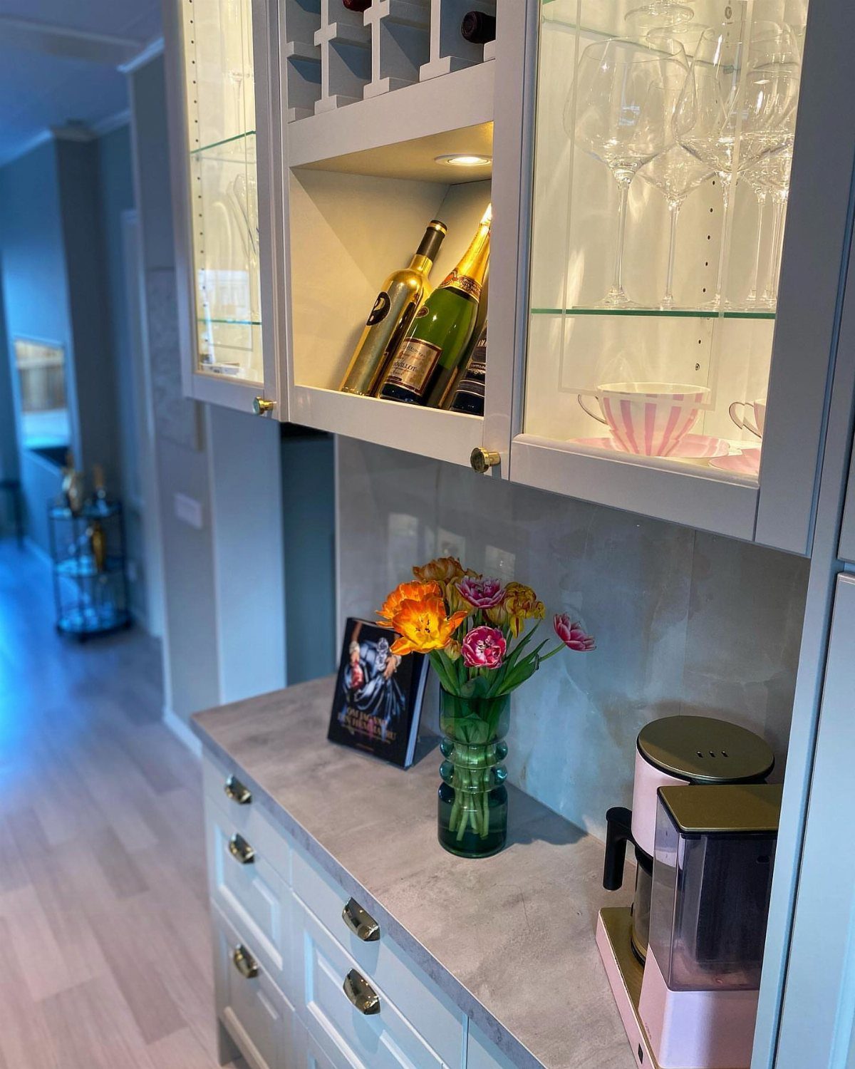 White kitchen cabinets containing wine bottles, glassware, and a pink coffee machine on a marble counter.