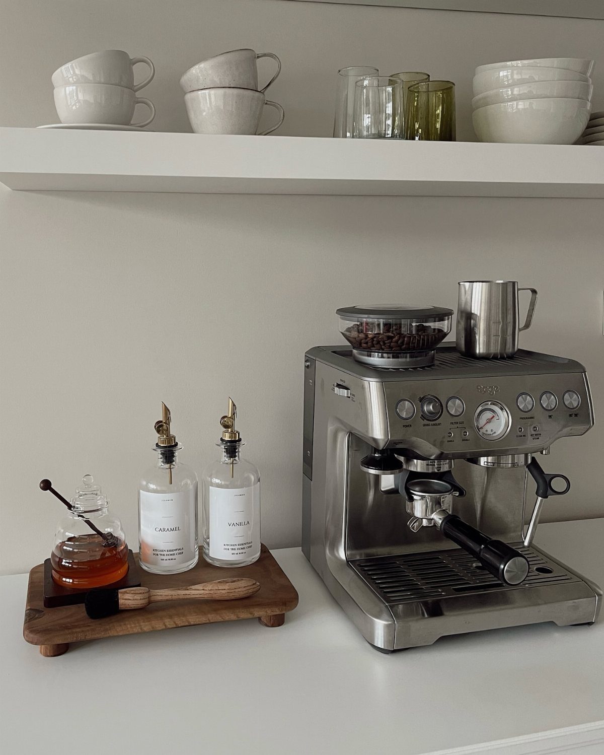 Silver espresso machine and syrup bottles on a white countertop with white shelving above.