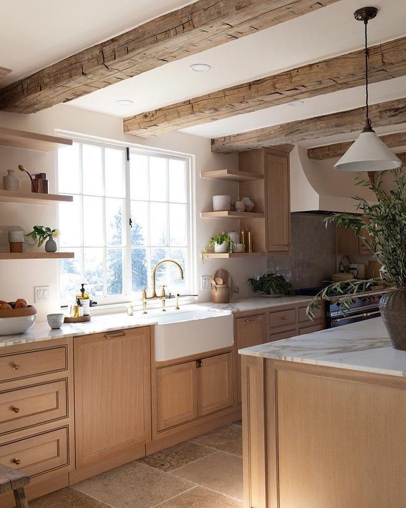 A bright kitchen with light wood cabinets, marble countertops, and large reclaimed wood ceiling beams.