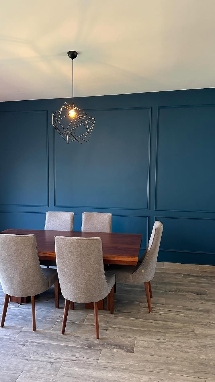 Grey upholstered chairs surround a wooden dining table against a dark blue paneled wall.