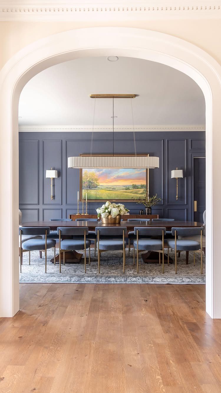 Dining room with dark blue paneled walls, blue chairs, and a large landscape painting under a gold chandelier.
