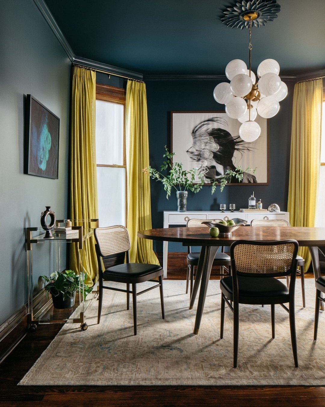 Dining room with dark blue walls, ceiling, and mustard yellow curtains surrounding a wooden table.