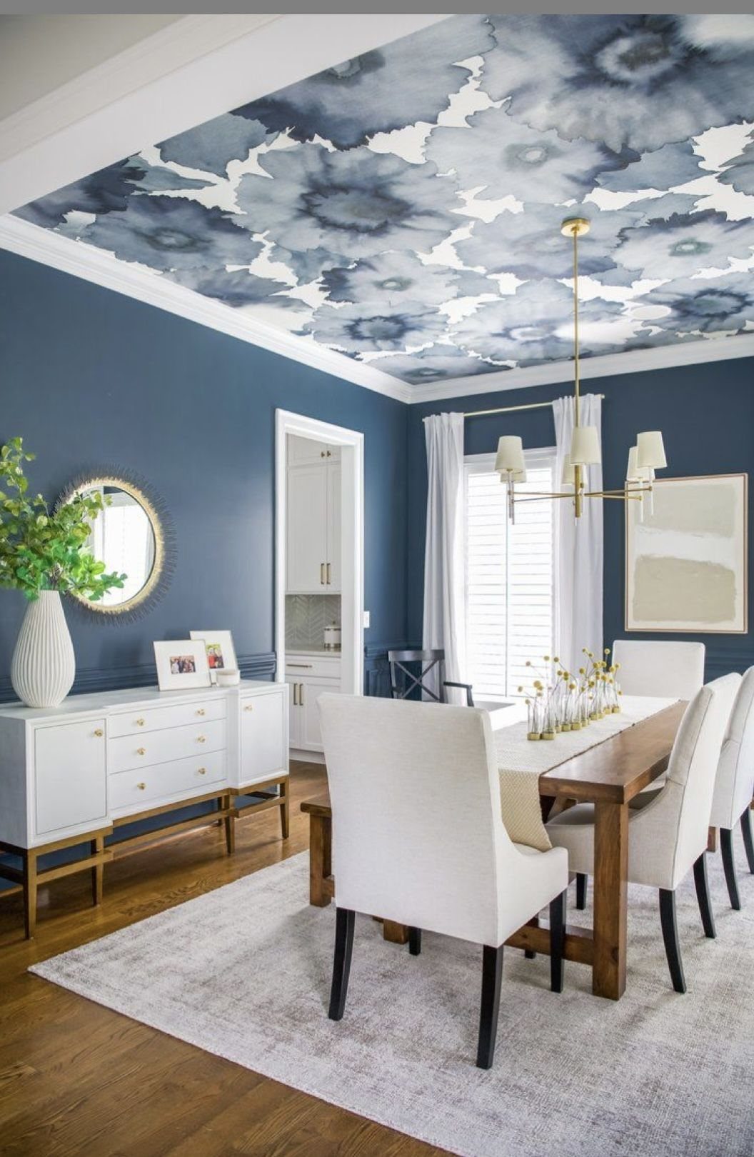 White upholstered chairs surround a wooden dining table in a blue room with a floral ceiling.