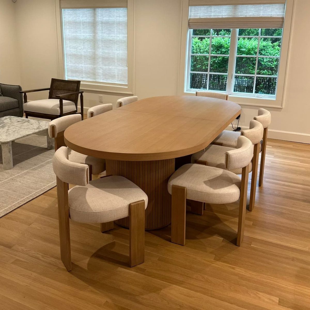A light wood oval dining table surrounded by six cream upholstered chairs in a bright room.