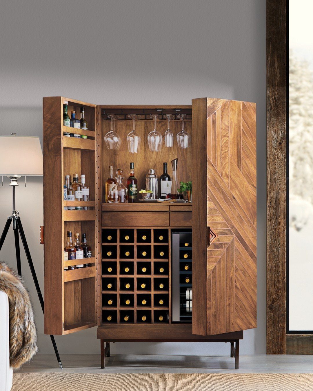 Wooden bar cabinet with open doors showing liquor bottles, wine glasses, and a wine cooler.