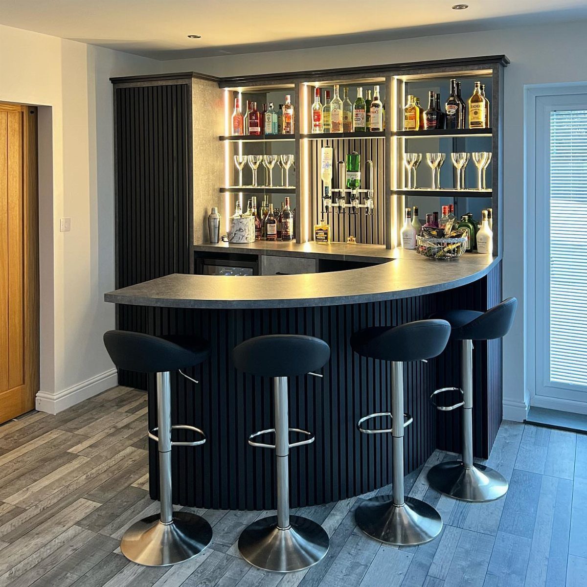 A curved dark wood home bar with illuminated shelves and four black bar stools.