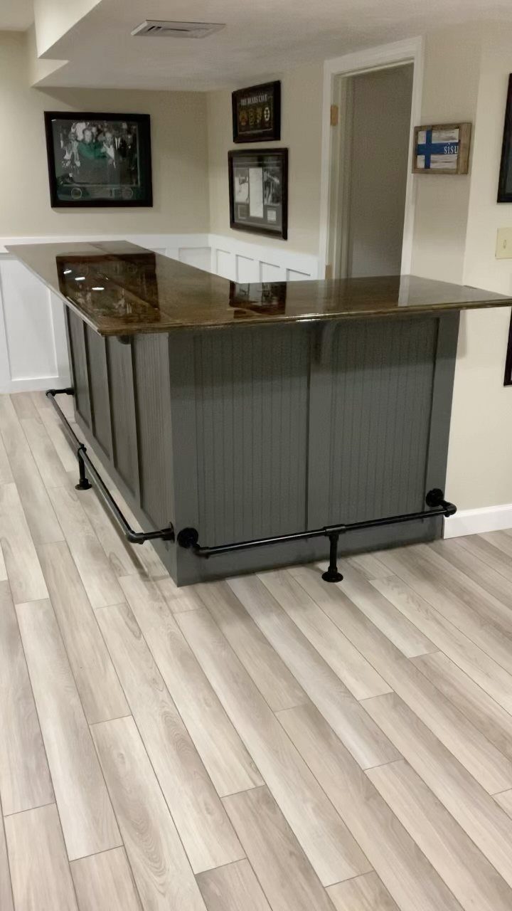 A grey paneled home bar with a polished dark countertop and black metal footrail.
